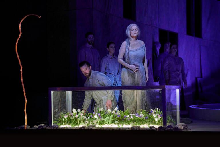 Erda stands before a glass case filled with flowers.